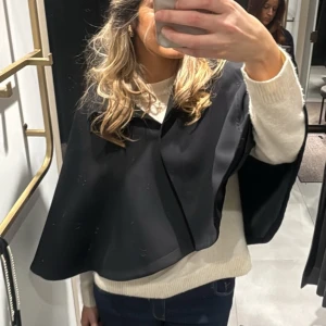 Svart cape-väst - Snygg svart cape-väst med en elegant och flytande design. Perfekt för att lägga till en stilfull touch till din outfit. Västen har en enkel och chic look som passar till många olika stilar.