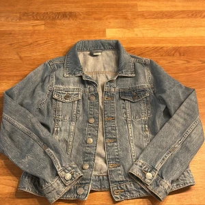 Jeansjacka från Lindex - Säljer en klassisk jeansjacka från Lindex i storlek 146. Jackan har en snygg blå tvätt och knappar framtill. Perfekt för vår och sommar!
