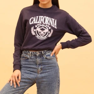 Mörkblå sweatshirt med tryck - Säljer en mörkblå sweatshirt med ett stort 'California' tryck i vitt och rosa på framsidan. Tröjan har långa ärmar och en rund halsringning. Perfekt för en avslappnad stil.