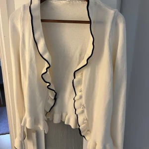 Vit kofta med volangdetaljer - Charmig vit kofta med svarta kanter och volangdetaljer. Den har knappar framtill och långa ärmar, perfekt för en stilfull look. Passar bra till både jeans och kjol.