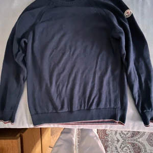 Mörkblå tröja från Moncler - Säljer en stilren mörkblå tröja från Moncler med rund hals och ribbade muddar. Tröjan har ett broderat Moncler-märke på ärmen och ränder i kragen. Perfekt för en avslappnad och snygg look. Tar emot byten 