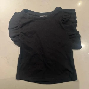 Svart ribbad topp från Zara - Säljer en svart ribbad topp från Zara med volangdetaljer på ärmarna. Toppen har en rund halsringning och korta ärmar.