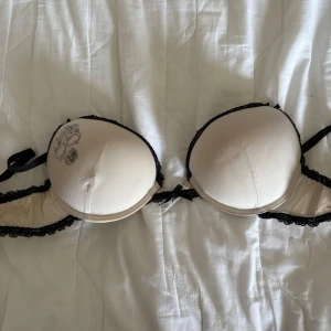 Beige och svart super Pusch up bh med dödskallemotiv - Snygg beige super push up bh med svart spetsdetalj och ett coolt dödskallemotiv på kupan. Den har en liten rosett framtill och justerbara axelband. Perfekt för dig som vill ha något unikt och stilfullt. Väl använd, där rosetten sitter mellan där så är den sliten. Står inte storlek men gissar på 75A-B