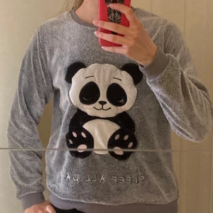 Grå mjukis tröja med panda - Mysig grå mjukis tröja med en söt panda-applikation på framsidan. Tröjan har långa ärmar och ribbade muddar vid ärmslut och nederkant. Perfekt för en avslappnad dag hemma.