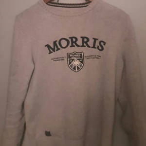Grå tröja från Morris - Säljer en stilren grå tröja från Morris med svart text och emblem på framsidan. Tröjan har långa ärmar och en rund halsringning. Perfekt för en avslappnad och snygg look.
