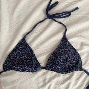 Blommig bikini med knytband - Söt bikini med blommigt mönster i rosa och grönt på en mörkblå bas. Toppen har trekantsform och knytband runt nacken och ryggen. Bikinitrosan har knytband på sidorna för justerbar passform.