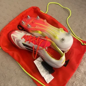 Adidas F50 Elite SG fotbollsskor - Säljer ett par oanvända Adidas F50 Elite SG fotbollsskor i vitt med röda och svarta detaljer. Skorna har snörning och är designade för optimal prestanda på planen med sina metallskruvdobbar. Perfekta för fotbollsspelare som söker både stil och funktion. Storlek 43