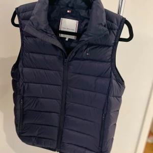 Mörkblå dunväst från Tommy Hilfiger - Snygg mörkblå dunväst från Tommy Hilfiger med dragkedja framtill! Den är i färgen marinblå och passar perfekt till allt! Den är perfekt nu till våren och sommaren! 