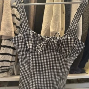 Rutig svartvit linnetopp med snörning - Supersöt linnetopp i svartvitt rutigt mönster med smala axelband och snörning framtill vid bysten. Figurnära passform som ger en snygg siluett, perfekt till jeans eller kjol. Toppens material känns lätt och passar grymt till varmare dagar.