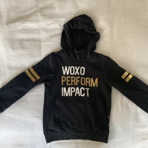 Svart hoodie från WOXO med guldtryck - Svart hoodie från WOXO med texten 'WOXO PERFORM IMPACT' i vitt och guld framtill. Guldiga ränder på ärmarna, dragkedjefickor och huva. Perfekt för dig som gillar sportig och cool stil.