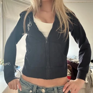 Svart velour hoodie från Svea - Svart croppad hoodie från Svea i mjuk velour med dragkedja framtill och huva. Broderad Svea-logga på bröstet och ärmen. Perfekt för en chill och sportig look.