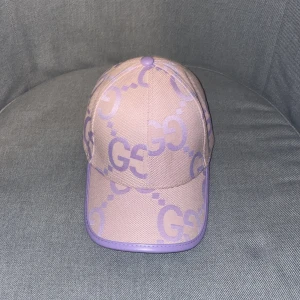 Ljuslila Gucci keps - Snygg keps från Gucci i ljuslila med stora GG-loggor i lila över hela kepsen. Kepsen har böjd skärm och lila kantband. Tillverkad i exklusiv merinoull och kashmir för en lyxig känsla. Perfekt för dig som vill sticka ut med en trendig accessoar. Denna keps är väldigt sällsynt och svår att hitta. 