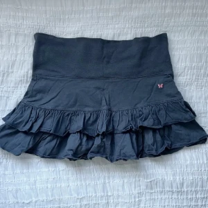 Mörkblå volangkjol med fjäril - Supersöt mörkblå kortkjol med dubbla volanger och bred midja. Liten rosa fjäril broderad framtill som extra detalj. Kjolen är gjord i mjuk bomull och har en avslappnad, flowy vibe som är perfekt för en chill look.