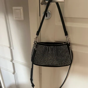 Svart axelväska med silvernitar - Cool svart axelväska från Zara  med massor av silverfärgade nitar på framsidan. Väskan har justerbar axelrem, dragkedja upptill och snygga metalldetaljer. Perfekt för dig som vill sticka ut med en edgy accessoar.