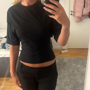 Svart topp med knyt i ryggen - Säljer en svart croppad topp med korta ärmar och snygg öppen rygg som knyts med en rosett baktill. Toppen har en enkel och stilren design, perfekt för dig som gillar trendiga detaljer och vill visa lite hud.