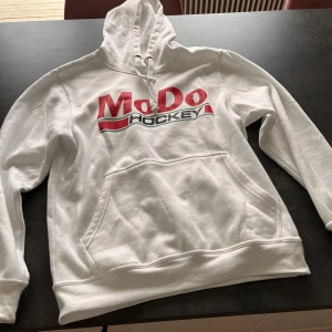 Vit MODO Hockey hoodie från Clique - Vit hoodie från Clique i storlek M med MODO Hockey-tryck i rött och svart framtill. Klassisk känguruficka, snörning i huvan och mjukt material. Perfekt för dig som vill visa ditt hockeyintresse och hålla stilen. Aldrig använd.
