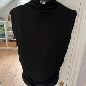 Svart kortärmad topp med scrunch i midjan - Svart ärmlös topp från H&M med hög hals och snygg rynkdetalj på sidan. Toppens material är mjukt och stretchigt, vilket gör den bekväm och enkel att styla. Perfekt för dig som gillar minimalistisk och stilren look.