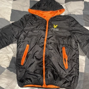 Svart pufferjacka från Lyle & Scott - Svart pufferjacka från Lyle & Scott Golf med orange foder och detaljer. Jackan har huva, dragkedja och två fickor framtill. Materialet är glansig polyester och jackan har en gul logga på bröstet. Perfekt för kyliga dagar när du vill sticka ut lite extra.
