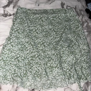 Grön blommig omlottkjol - Supersöt grön kort omlottkjol med vitt blommönster. Kjolen har volangkant och knyts i sidan för en extra snygg detalj. Perfekt för dig som gillar en fräsch och somrig vibe.