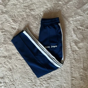 Palm Angels blå träningsbyxor - Mörkblå träningsbyxor från Palm Angels med vita sidoränder och broderad logga på låret. Byxorna har elastisk midja och dragkedjor vid bensluten. Snygg och sportig look som passar perfekt till streetwear.