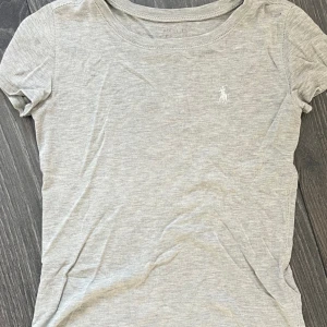 Grå t-shirt från Ralph Lauren - Snygg ljusgrå t-shirt från Ralph Lauren, passar till allt och så skön att ha på sig. Väl använd men fortfarande fint skick! Inga fläckar 💞 tvättas och styrks innan köp💞 köptes för länge sedan men passar nog 140, kan skicka fler bilder🩷 jag är stl 146-152 och kan ha den men den är lite kort!