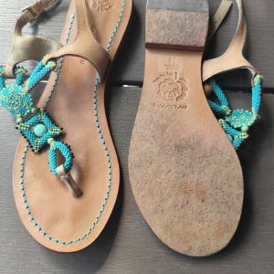 Beiga sandaler med turkosa pärlor - Snygga sandaler från Maliparmi i beige skinn med handgjorda pärldetaljer i turkost, blått och grönt. Tåremmen är dekorerad med pärlor i olika nyanser och mönster, vilket ger en bohemisk och unik look. Perfekta för sommardagar och festivaler.