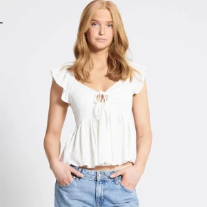 Vit blus med volang och knyt - Supersöt vit blus med volangärmar och knyt fram. Den har v-ringning, lätt rynkad struktur och är croppad med luftig passform. Perfekt för varma dagar.