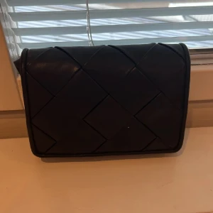 Svart axelväska i skinn från H&M - Säljer en svart axelväska från H&M med flätad design och stilren rektangulär form. Väskan har justerbar axelrem och är gjord i skinnimitation. Perfekt för dig som gillar minimalistisk och trendig look.