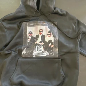 Svart ODZ hoodie med tryck - Svart hoodie från ODZ med stort färgglatt tryck på bröstet där det står 'ODZ Skator & Kråkor'. Tröjan har huva med snörning och en klassisk magficka. Perfekt för dig som gillar svensk hiphop och vill sticka ut med en unik streetstyle. Storlek xs/s