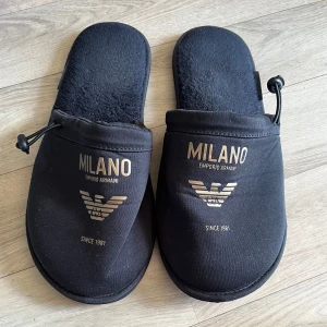 Svarta tofflor Emporio Armani Milano - Svarta tofflor från Emporio Armani Milano med guldig logga och text framtill. Mjukt foder och justerbar dragsko vid öppningen. Perfekta för chill hemma, med stilren design och bekväm passform.