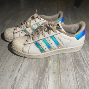 Adidas Superstar med holografiska detaljer - Säljer ett par vita Adidas Superstar sneakers med holografiska regnbågsfärgade ränder och häl. Klassisk snäcktå, snörning och platt sula. Skorna är i skinn och har den ikoniska Adidas-loggan på hälen och tungan. Perfekt för dig som vill sticka ut med coola detaljer.