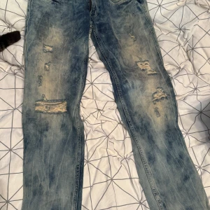 Blå slitna jeans från Replay - Säljer ett par blå jeans från Replay med slitna detaljer och hål framtill. Jeansen har en tvättad look med ljusa partier och raka ben. Klassisk femficksmodell och tydliga sömmar på bakfickorna. Perfekta för en avslappnad streetstil. S