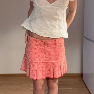 Lågmidjad Rosa blommig kortkjol med volang - Superfin rosa kortkjol med blommigt mönster och volang nertill. Kjolen har fickor fram och knäppning med knapp och dragkedja. Midjemått: 40cm, Längd: 33cm Storlek: M. Från Pepe jeans