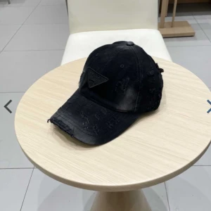 Svart distressed keps från Prada - Svart keps från Prada med coolt distressed och slitet utseende. Klassisk böjd skärm och justerbar baksida. Kepsen har en svart triangel-logga framtill och är tillverkad i bomull. Perfekt accessoar för en edgy streetstyle.