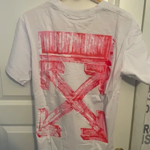 Vit Off-White t-shirt med rosa print - Snygg vit t-shirt från Off-White med ikoniskt rosa grafiskt tryck på ryggen och liten logga framtill. Klassisk passform med rund hals och korta ärmar. Perfekt för dig som vill sticka ut med streetwear-vibe.