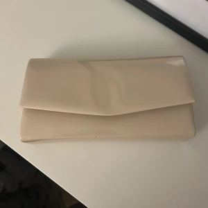 Beige kuvertväska i blankt material - Säljer en stilren beige kuvertväska med blank finish och enkel design. Väskan har ett innerfack och en avtagbar silverfärgad kedja så du kan bära den både i handen eller över axeln. Perfekt accessoar för bal eller liknande.