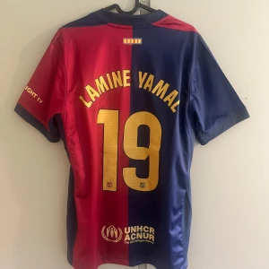 FC Barcelona Lamine Yamal matchtröja - Snygg FC Barcelona matchtröja med Lamine Yamal och nummer 19 på ryggen. Tröjan har klassiska blå och röda paneler, guldiga detaljer och klubbmärke på bröstet. Tillverkad i lätt och ventilerande polyester, perfekt för fotbollsälskare.