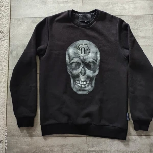 Svart sweatshirt från Philipp Plein - Svart sweatshirt från Philipp Plein med broderad logga och texten 'PHILIPP PLEIN 1978' på ryggen. Tröjan har rund hals, långa ärmar och ribbade muddar. Snygg detalj framtill med ett emblem. Perfekt för dig som gillar streetwear.