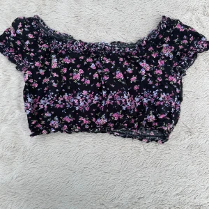 Svart blommig croppad topp - Supersöt croppad topp med svart botten och små blommor i rosa, lila och blått. Offshoulder-modell med korta ärmar och smockdetaljer vid halsringning och nederkant. Perfekt för en somrig look och lätt att matcha med jeans eller kjol.