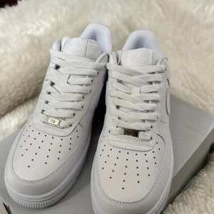 Nike Air Force 1 vita sneakers - Klassiska Nike Air Force 1 sneakers i helvitt skinn med perforerad tå, rund tå och platt sula. Snygg och tidlös modell med snörning och diskret Nike-logga på sidan. Perfekta för dig som gillar en clean och fräsch look.