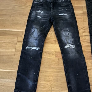 Svarta slitna jeans med raka ben - Snygga svarta jeans med slitningar och hål på knäna samt coola detaljer som ger en streetkänsla. Raka ben och klassisk femficksmodell. Perfekta för dig som gillar en edgy stil.