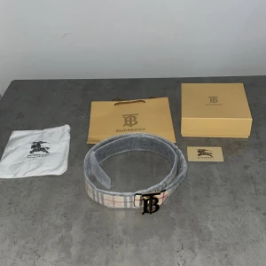 Burberry Rutig Bälte - Snyggt Burberry-bälte i klassiskt rutigt mönster med beige, svart, vit och röd färg. Bältet har ett ikoniskt TB-spänne i guld och är tillverkat i skinn. Insidan är svart och bältet levereras med originalpåse, ask och kort. Perfekt accessoar för att lyfta din outfit. Varan fraktas ut dagen du beställer eller dagen efter. STL 110cm
