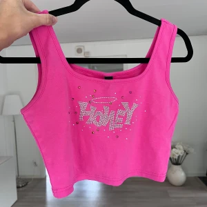 Rosa croppad topp med strass - Superfin croppad topp med strass i stark rosa färg. Framsidan har texten 'HONEY' i glittriga stenar och små färgglada pärlor runtom. Topp med breda axelband och stretchigt material! Önskar du veta exakta mått? Tveka inte att höra av dig! ALDRIG ANVÄND