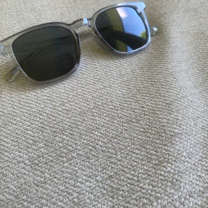 Grå transparenta solglasögon - Snygga solglasögon med transparent grå båge och svarta fyrkantiga glas. Ramen är i plast och har en modern, stilren look som passar perfekt till sommaren. Enkla att matcha med olika outfits och ger en cool vibe.