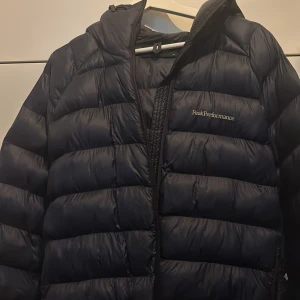 Svart dunjacka Peak Performance - Snygg svart dunjacka från Peak Performance med broderad logga på bröstet och ärmen. Jackan har huva, dragkedja och klassisk pufferstil med breda kanaler. Perfekt för kalla dagar och riktigt skön att bära.