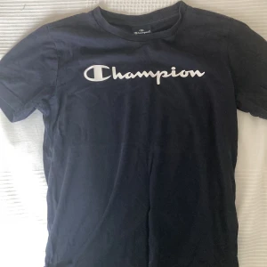 Mörkblå Champion t-shirt i bomull - Snygg mörkblå t-shirt från Champion med klassisk logga i vitt framtill och liten logga på ärmen. T-shirten är i mjuk bomull och har en normal passform med korta ärmar. Perfekt för en chill och sportig look.