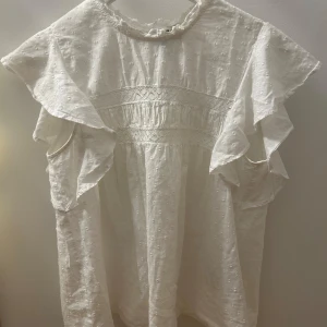 Vit blus med volang från Vero Moda - Supersöt vit blus från Vero Moda med volangärmar och små broderade prickar i tyget. Blusen har en hög hals med liten volangkant och fina spetsdetaljer över bröstet.