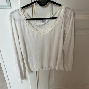 Vit långärmad topp med spets  - Supersöt vit långärmad topp från Amisu med ribbad struktur och v-ringning dekorerad med spets. Lätt croppad känsla med vågig kant nertill och vid ärmslut. Perfekt för lager-på-lager eller som den är till jeans eller kjol.