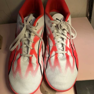 Puma vita och röda fotbollsskor - Snygga fotbollsskor från Puma i storlek 46.5. Skorna är vita med röda och svarta detaljer, har snörning och en platt, röd sula som passar perfekt för inomhusspel. Ovandelen är i syntetmaterial och designen är sportig och modern.