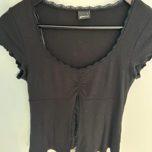 Svart topp med spets från Gina Tricot - Svart kortärmad topp från Gina Tricot med v-ringning och spetsdetaljer längs halsringningen och framtill. Toppen har en liten slits framtill och rynk vid bysten. Perfekt för dig som gillar en feminin och trendig stil.